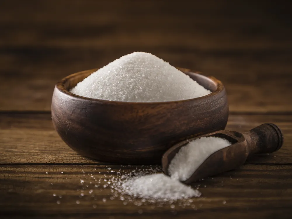 White sugar in a brown wooden bowl with a serving scoop in front with sugar pouring out