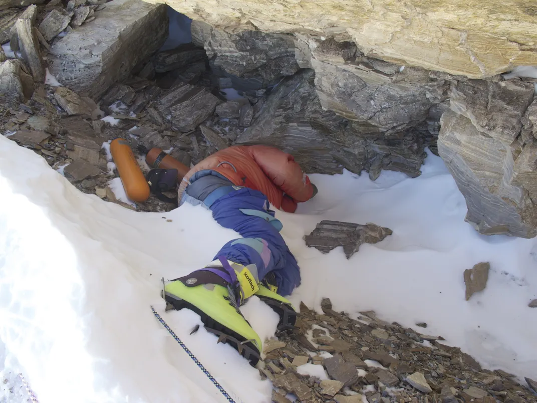 body of a mountaineer in winter gear partially covered in snow with rocks nearby. They are wearing green climbing boots